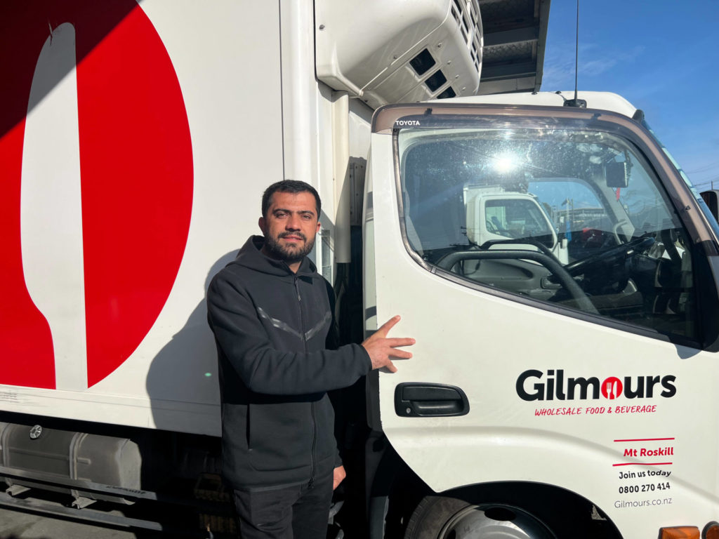 Picture of Afghan refugee Akbar Hashimi next to a Gilmours Food and Bervrage Wholesalers vehicel