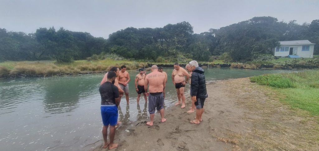 Men stand in a circle next to water for Karakia  on Rangitoto Island