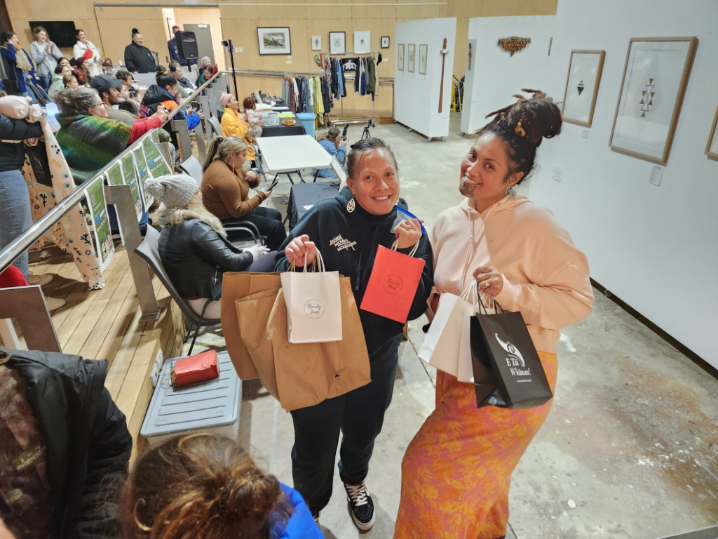 Two wāhine (women) smile and display E Tū Whānau bags and other goodies at the Mamas Moving Mountains Matariki 2023 event in