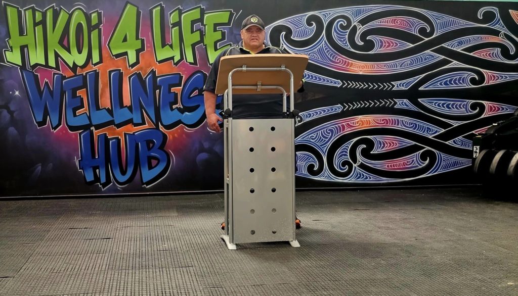 Les Hokianga stands at a lectern speaking at the opening of the Hub. Behind him a mural says "Hikoi4Life Wellness Hub".