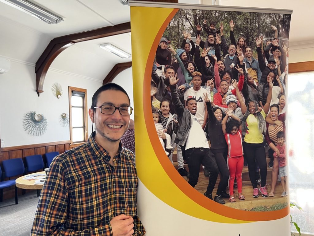 Sergio Opazo, Escular del Buen Vivir Programme Director for the ALAC Wellbeing Toolbox smiles next to a banner showing a crowd of smiling people.