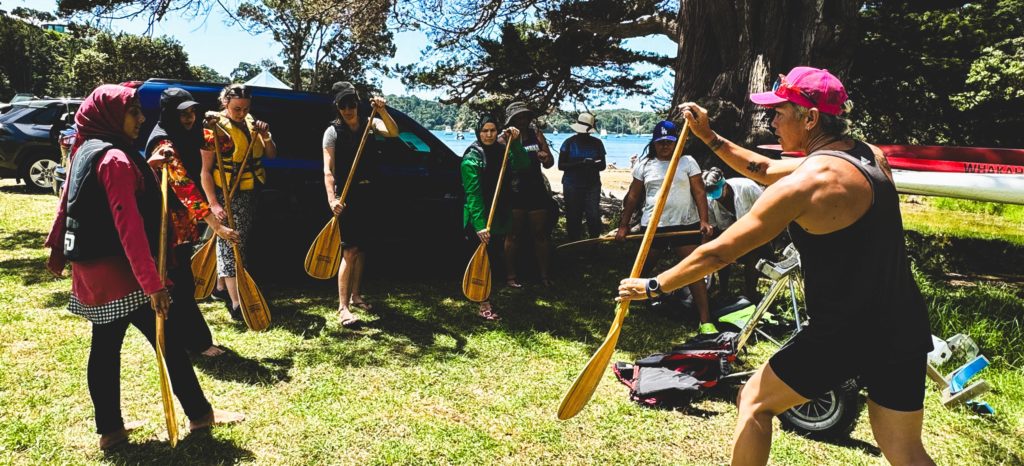 The NFACT whānau enjoyed learning waka ama from the wāhine from Māma Moving Mountains. Doing so offered plenty of rich connections and conversations about te ao Māori and life in New Zealand. In this photo, one woman teaches a group of rangatahi how to use a paddle. They're all wearing life jackets and are standing in a semi circle under a tree with the sea in the background.