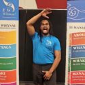 At the National Māori Basketball Tournament 2024, E Tū Whānau kaimahi Mōwai Smiler performs a pūkana with eyes wide open and hands raised between two banners showing the six E Tū Whānau values. These are Aroha, Whanaungatanga, Whakapapa, Mana manaaki, Kōrero awhi, Tikanga.