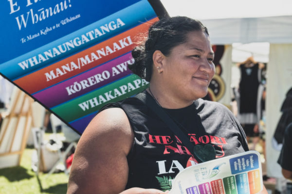 A wahine holds an E Tū Whānau values fan and walks in front of a banner dispalying the E Tū Whānau values at Waitangi 2024.