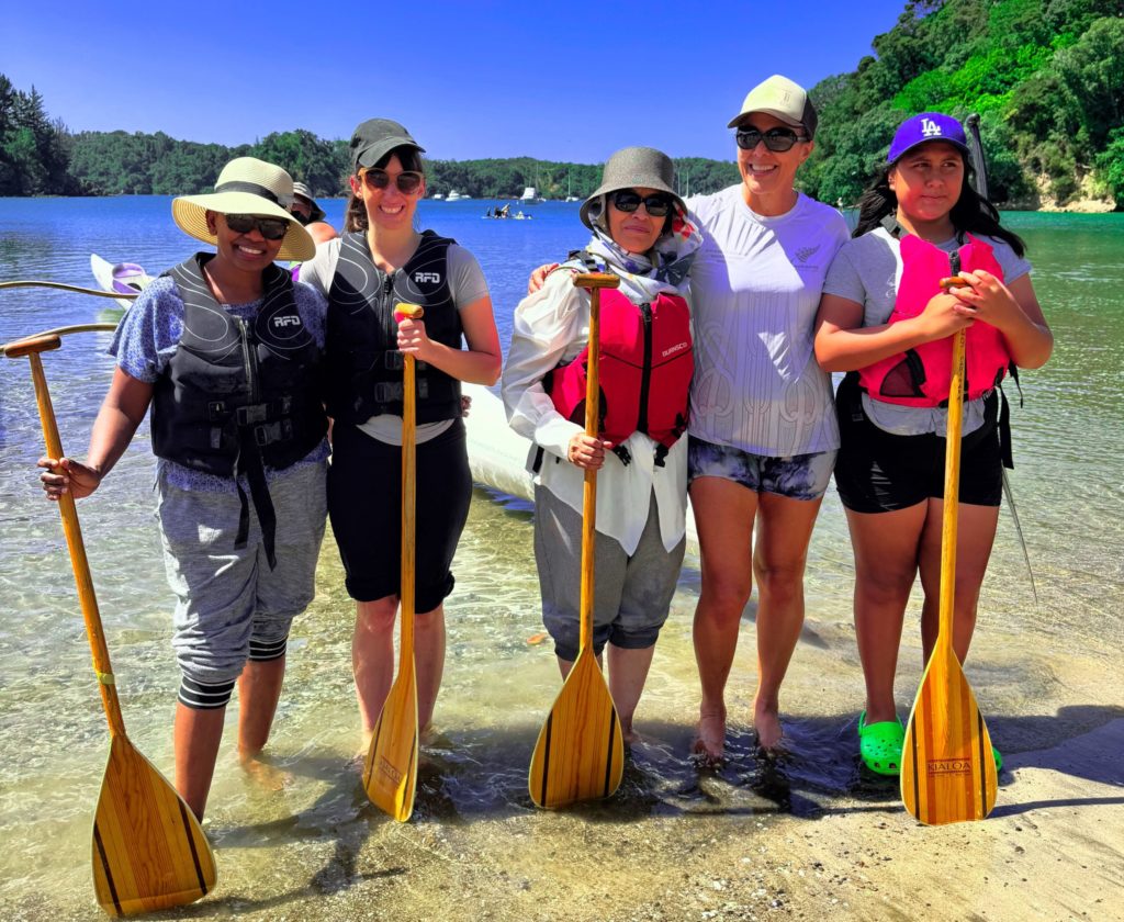 Some of the NFACT rangatahi and wāhine from Māma Moving Mountains enjoyed rich connections while learning waka ama. Here fice of them stand in ankle deep water. They're all smiling as they pose for a photo together.