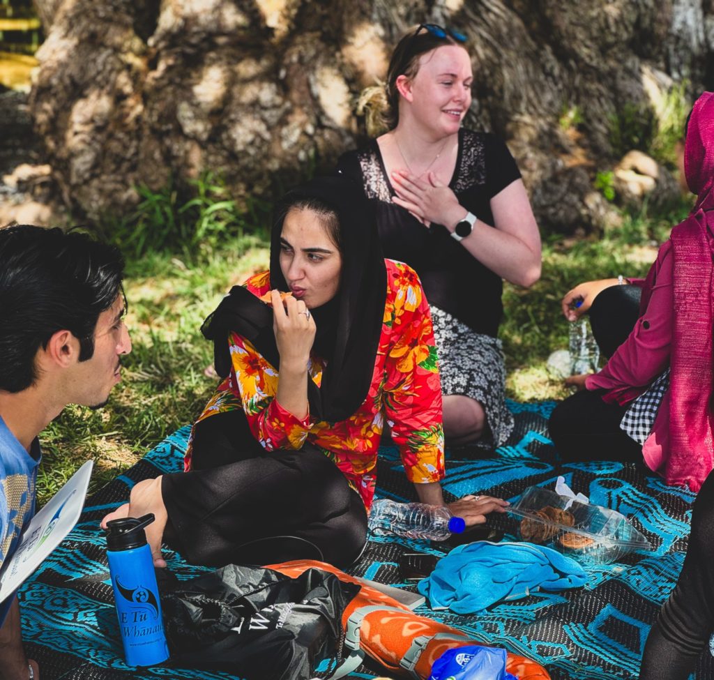 The experience of Waitangi 2024 included eating together. Int his photo, people sit and chat on bright coloured rugs. One woman who wears a Muslim headscarf is eating as she talks to a young man. the scene is colourful and bright. 