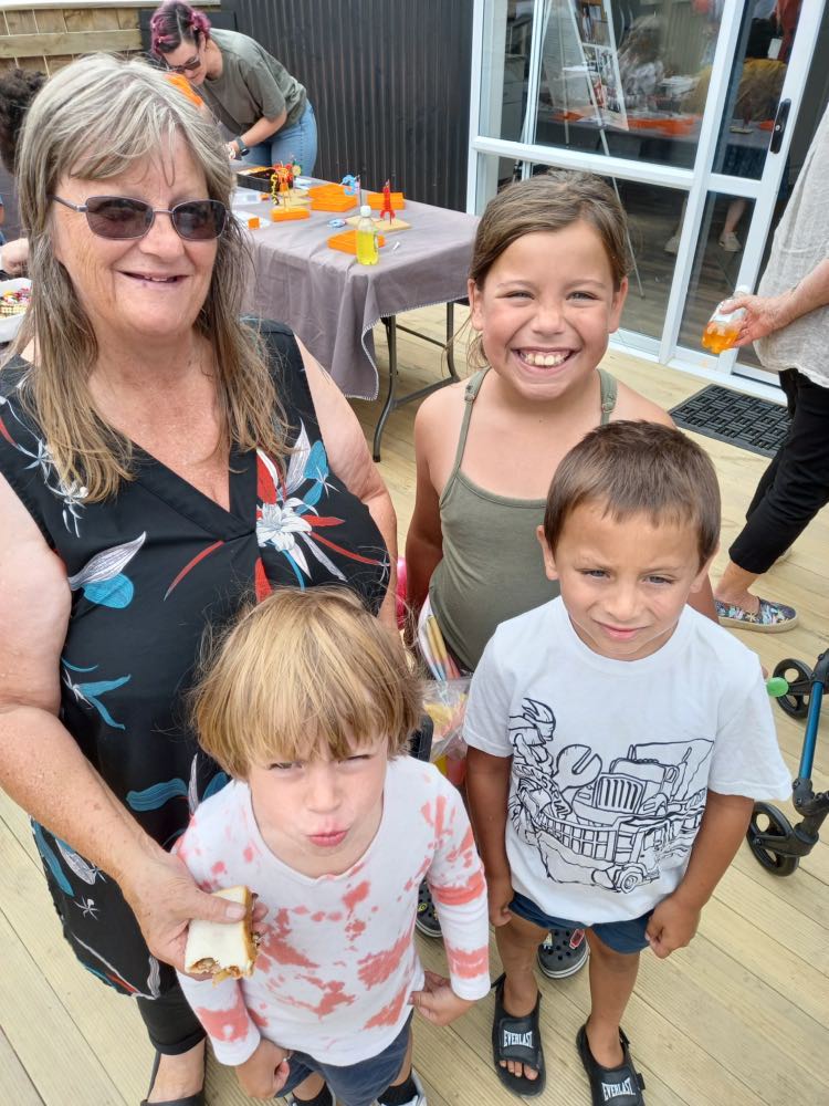 This image is of Local resident Deborah Poulton with her three young moko at the Tuia te Hāpori opening day. Deborah is holding some food. Behind the group, a table with BBQ food is visible.