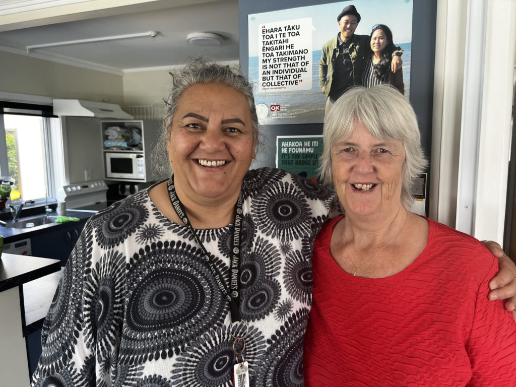 Street Co-ordinator, Donna McKinnon and her ever supportive colleague, Robyn Parkes, Whakatu Te Korowai Manaakitangi Trust Manager stand with arms around each other smiling at the camera. They are in a kitchen. Behind them, a poster says "Ehara tāku toa i te toa takitahi ēngari he toa takimano. My strength is not that of an individual but that of a collective."