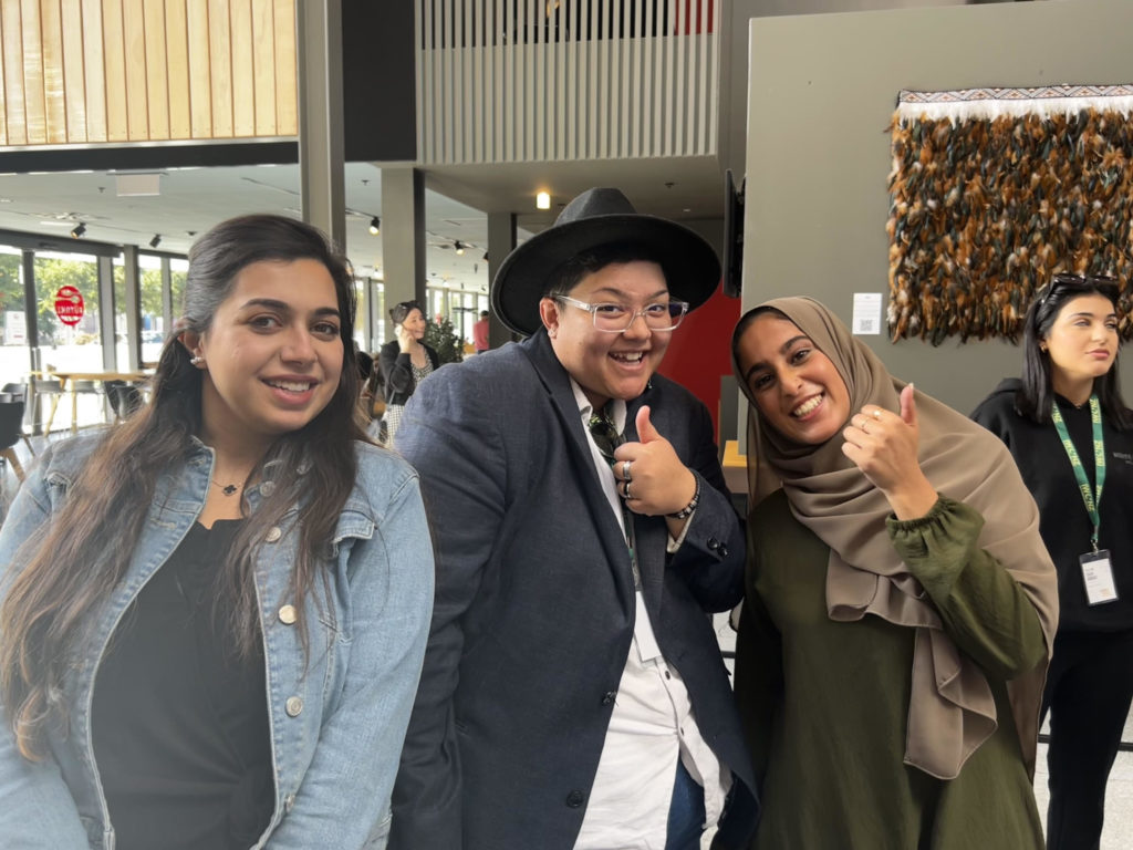 ICWNZ National Conference 2024 organisers Rand Alomar (Left) and Nabeela Ahmed (Right) smile for a photo with Shaneece Brunning, E Tū Whānau Rangatahi Coordinator. They are in the lobby off the conference venue, and Shaneece and Nabeela are hold their thumbs up while all three women smile at the camera.