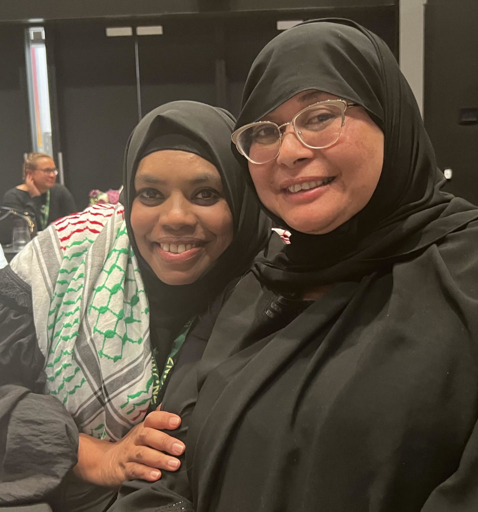 Two of the participants at ICWNZ National Conference 2024, Farina Khan and Miriam Moton, look at the camera and smile in this photo. Both women are wearing hijab. 