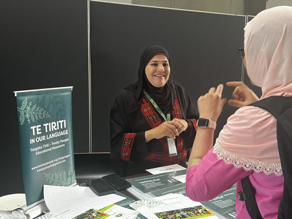 Participants at IWCNZ National Conference 2024 were able to engage with Te Tiriti o Waitangi. In this photo, two women wearing hijab talk to each other. Between them on a table are various documents and small banner which says: "Te Tiriti in our language. Tangata Tiriti - Treaty People Education Resources".