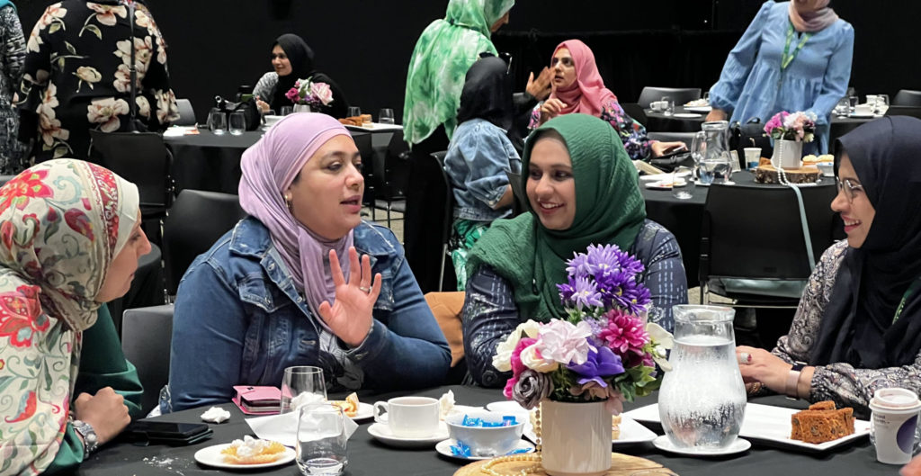 This photo is of three tables of women who are talking together at ICWNZ National Conference 2024.  In the foreground of the picture,  four women, all wearing hijab, look relaxed and happy as they talk. 