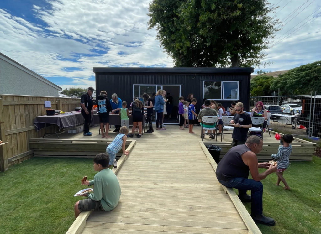 This image is of a group of people sitting along a wooden walk way and deck outside a small black building at the opening day barbeque for Tuia te Hāpori. There are about 30 people eating together on a warm-looking day.