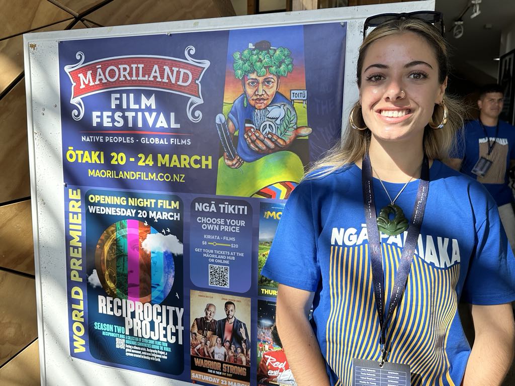 In this picture, Oriwa Hakaraia, Pou Ruruku Rangatahi at Māoriland, and coordinator of E Tū Whānau Filmmaking Workshops smiles as she stands next to big poster. The poster is bright and colourful and is for Māoriland Film Festival 2024.