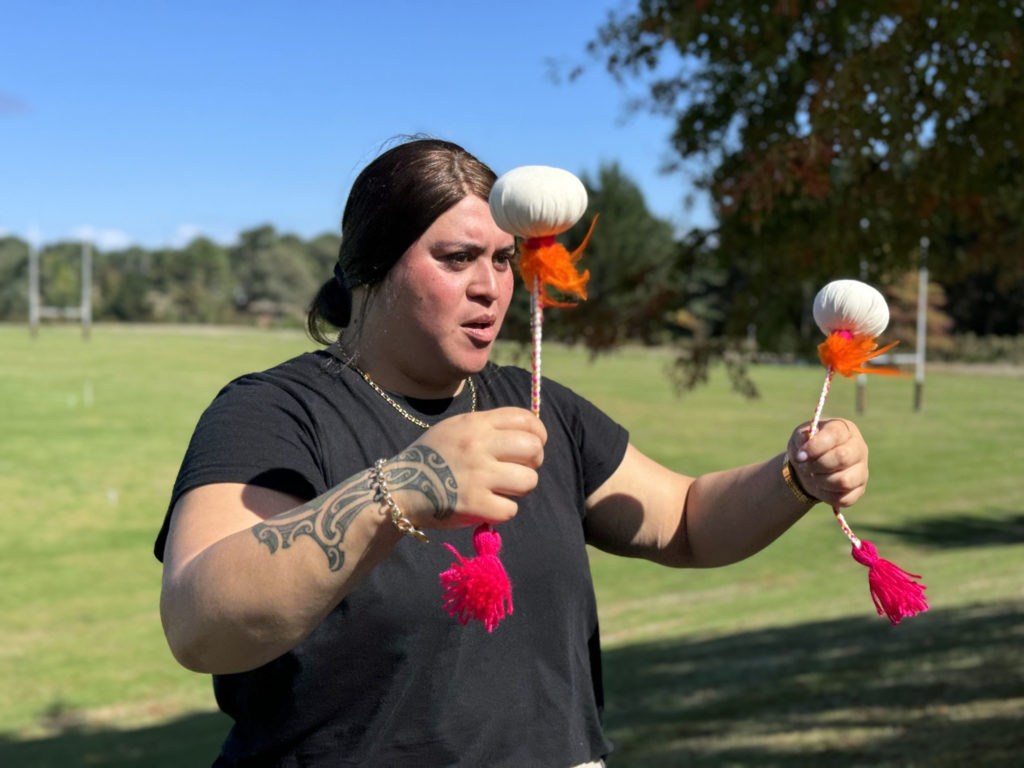 A woman is pictured doing spin poi at Te Hapori Ora POD. She has a tattoo on her right forearm, which closest to camera. Both arms are raised and she looks in concentration as she spins a poi in each hand. The poi are white with orange feathers attaching them to a multi-coloured string, at the end of which are fluffy red ties. She is outside on a blue, sunny day.