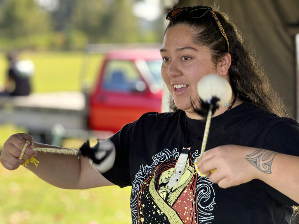 This close up is of a young woman whose face is bright and open as she spins poi at Te Hapori Ora POD. The poi are in movement and are white with black feathers attaching the hand-held cord. She wears a dark coloured t shirt with an ethnic print on the front. The background is blurred.