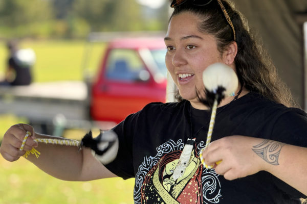 This close up is of a young woman whose face is bright and open as she spins poi. The poi are in movement and are white with black feathers attaching the hand-held cord. She wears a dark coloured t shirt with an ethnic print on the front. The background is blurred.
