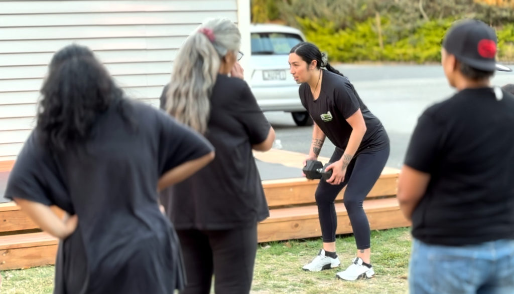This over the shoulder image, a woman is in a standing crouch as she holds a dumb bell in front of her with both hands. In the foreground of the image, three people are pictured from behind as they look at the woman who is completing the exercise circuit at Te Hapori Ora POD.