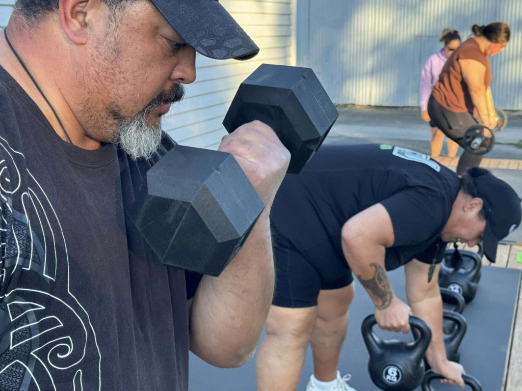This close up image is of a man doing arms curls with a dumb bell on a wooden deck outside the Te Hapori Ora POD. He is dressed in dark clothing and wears a hat. In the background, a woman bends over and lifts a kettlebell while in the background a woman lifts a barbell while another woman looks on.