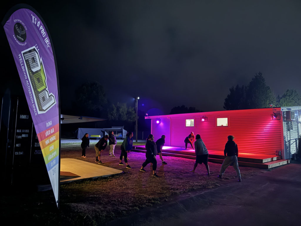 In this picture, people take part in a night time cardio and coordination session at Te Hapori Ora POD. They're outside and the building behind them is illuminated with pink and purple lights. In the foreground is a tear drop flag saying Te Hapori Ora POD - Wellbeing Activation Space.