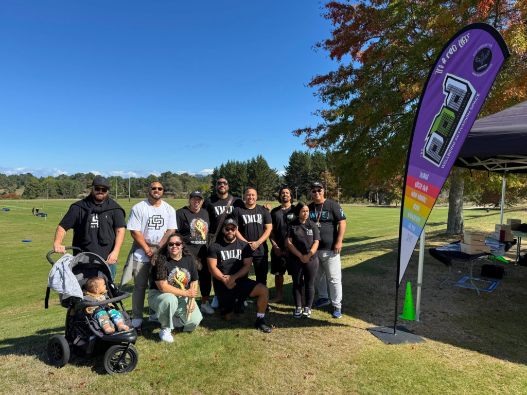 Whānau from Te Hapori Ora, including William Olds, pose for a photo. There are 11 people in total, arranged in a back standing row, with some people kneeling in front. Some are women but most are men. One man holds a pram with a baby sitting in it. Next to them is a colourful tear drop flag on which we can read POD Wellbeing Activation Space. Some other words are not legible.