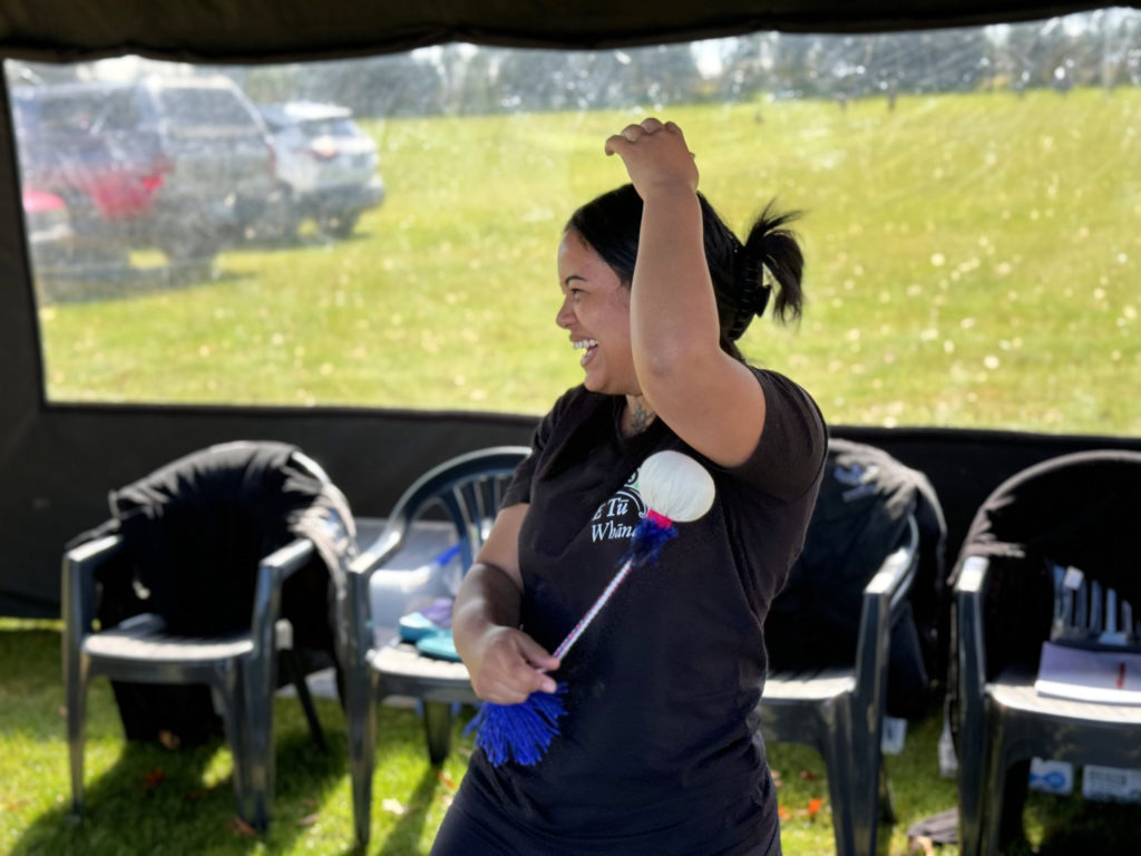 A woman wears a black t shirt showing most of the E Tū Whānau name and logo. She stands in some sort of tent at Te Hapori Ora POD, through which the day appears sunny. In her right hand, she spins a poi. Her left hand is raised above her head. She smiles and is looking out of the picture frame.