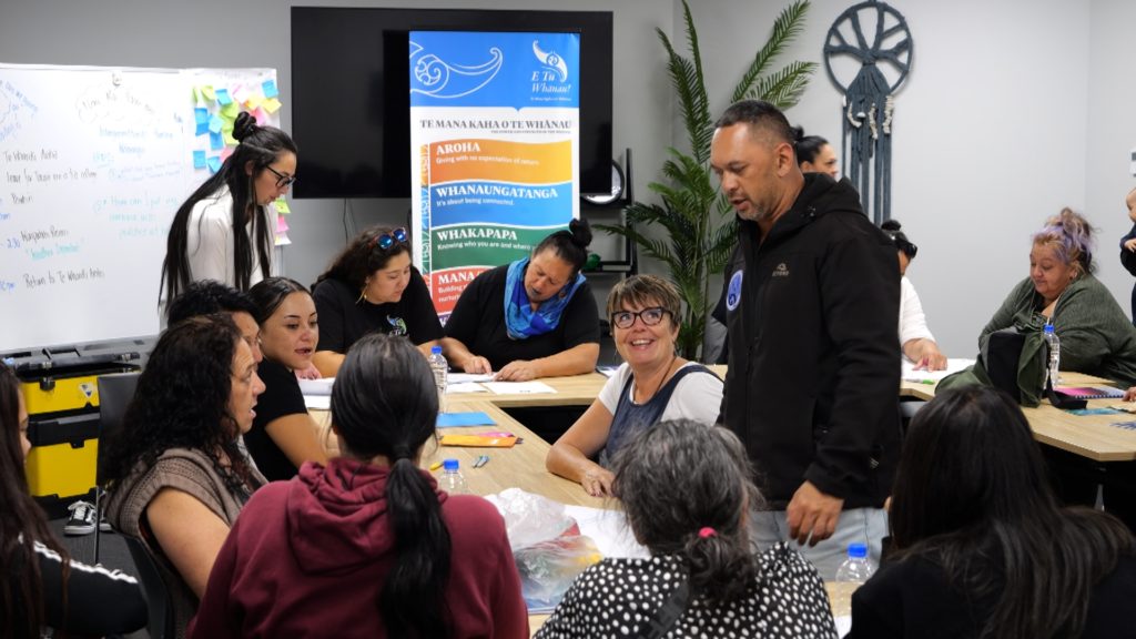 A group of adults sit and work together at a wellbeing wānanga run by Te Hapori Ora POD. IN the foreground, a a man is standing while others in the group who are sitting are looking at him and smiling. Papers and booklets cover the desk. In the background, is an E Tū Whānau flag and various people are sitting and working too. One woman is standing as she works.