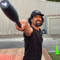 William Olds smiles as he looks at the camera. He has mid length hair and a goatee beard. In his right hand, swung up towards the viewer, is a long, black kettlebell. He is doing some sort of workout on a deck outside building.