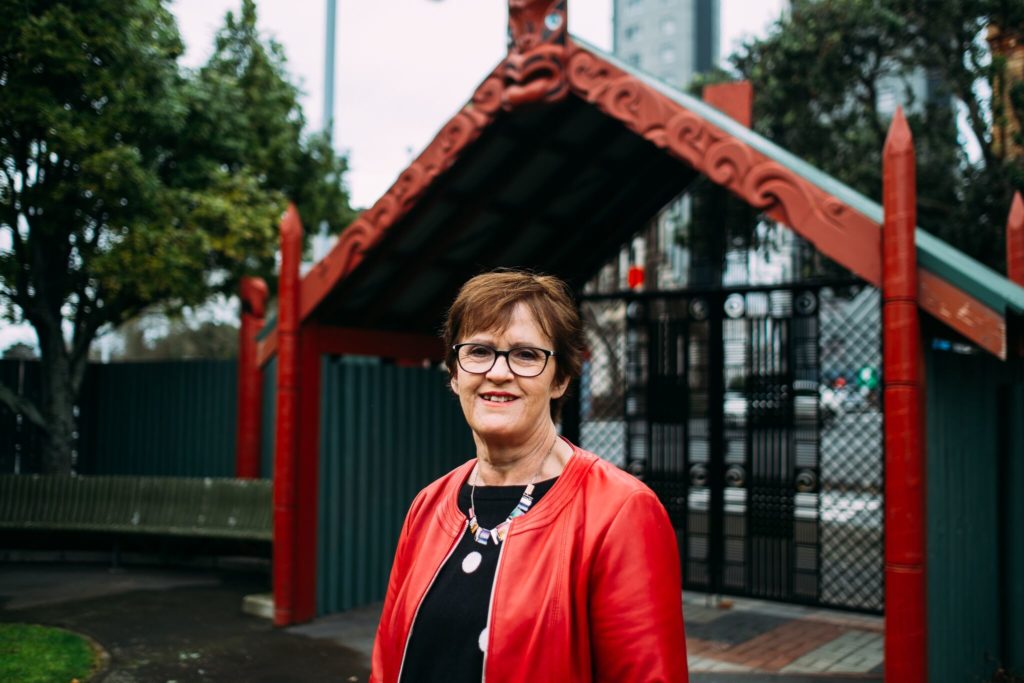 Densie Wilson, who led the research and review of contemporary Māori research on Māori living with violence and sexual violation is pictured. She has short hear, glasses and is smiling as she looks at the camera. She wears red and black clothing and bright coloured necklace and is standing in front of a waha roa or entry way to a marae.