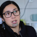 E Tū Whānau kahukura, Savanna Martin-Koteka, is pictured close up. She wears glasses and dark clothing. She has a moko kauae tattoo on her chin and a small silver ring in her left nostril. She is looking attentively past the camera.