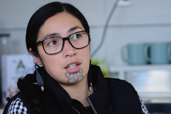 E Tū Whānau kahukura, Savanna Martin-Koteka, is pictured close up. She wears glasses and dark clothing. She has a moko kauae tattoo on her chin and a small silver ring in her left nostril. She is looking attentively past the camera.