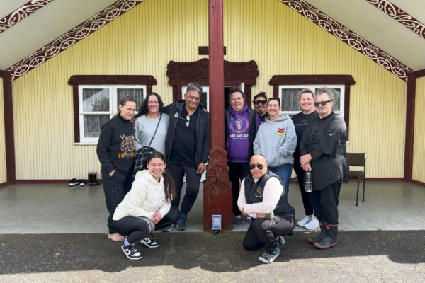 People who attended Te Korowai Aroha o Aotearoa wānanga stand together in front of the whare nui at Matau Marae. Ten people are pictured. They stannd close together, some with an arm around each other. They are all smiling.