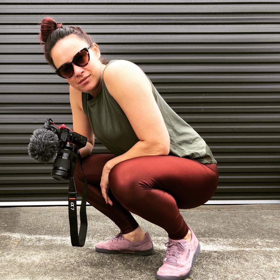 Aroha Treacher  of Mana Wāhine Hauora is pictured crouching on her haunches. She holds a camera and is wearing dark glasses, red leggings and a green singlet. She smiles and looks confidently at the camera.