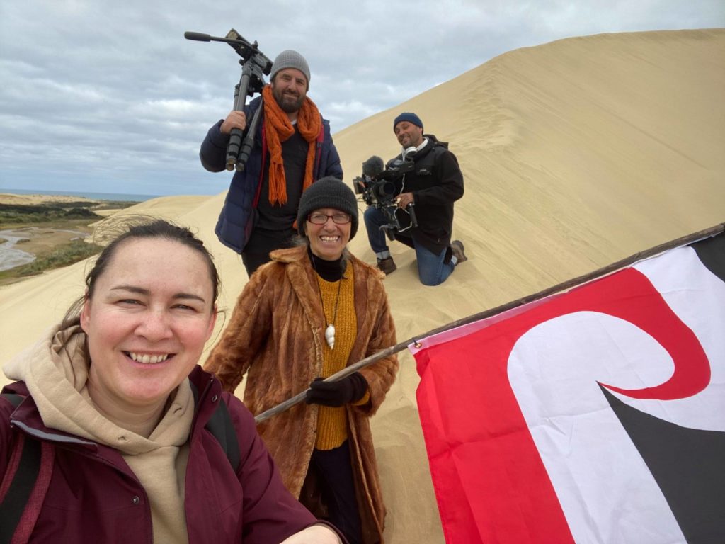 Aroha Treacher of Mana Wāhine Hauora is pictured on a beach dune holding a Tino Rangtiratanga flag. Behind her are three other people, all holding various types of filming equipment.