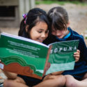 Two chldren sit next to each other on the ground outside. They are smiling and looking at the book Tūpuna!. The cover of the book is green and brown, with white writing.