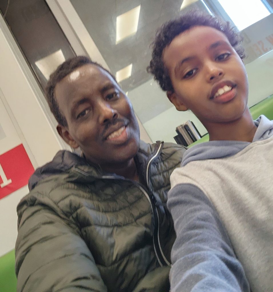 This selfie picture is of Abdirizak and his son Mohamad in an indoor setting. They both smile as they look down at the camera and are dressed in dark coloured casual clothing. Abdi, who is on the left is middle aged, while Mohamad appears to be aged about 10 years.