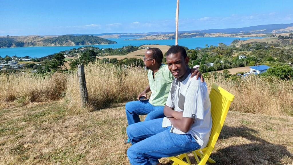 In this picture, two men who are wearing casual clothing, sit on a yellow chairs on a hill in a rural setting. The day is sunny and in the back ground, the ocean is visible. The man closest to the camera sits with his arms crossed and is looking at the ground. The second man has his arm across the shoulders of the first man.