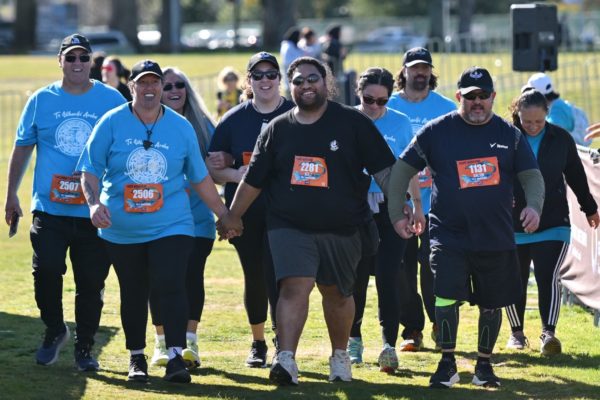 In this photo, a group of people cross the finish line of a marathon. They look happy and are all smiling. The three people in front are holding hands and close behind them, the rest of the group is visible.