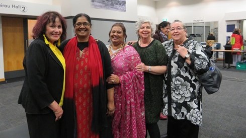 In this photograph, Margaret Western and four women of various ethnicities stand together in a line, Two of the women wear brightly coloured South Asian clothing. The women stand close together, with several with arms around each other or making other forms of friendly contact. They all look happy. The photograph is taken inside a large conference-type room.
