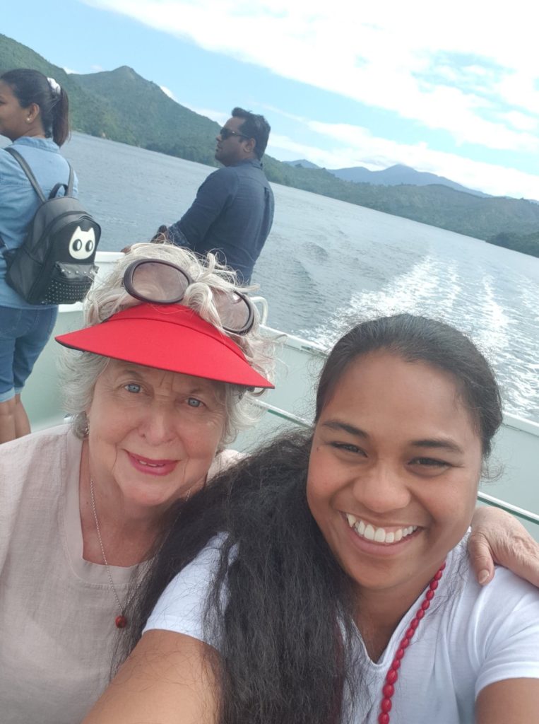 In this photograph taken on the back of a moving boat, Margaret Western wears a bright red cap and light coloured clothing. She has her arm around a young woman, Meeri, from Kiribati who is wearing a light coloured t shirt. Both women look happy and are looking directly at the camera. Two people are visible behind Margaret and Meeri on the boat and, behind them, the hills of the Marlborough Sounds can be seen.