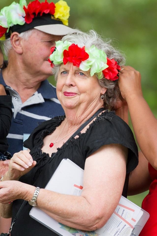 Margaret Western is seen in a mid-shot. She is an older woman, with kind blue eyes. Dressed in black, she holds an folder of papers under arm as someone, who is unseen apart from their hands, secures a garland of red and green flowers on her head. Behind Margaret, an older man wearing a blue and white stripped shirt and the same red and green garland of flowers, is visible.