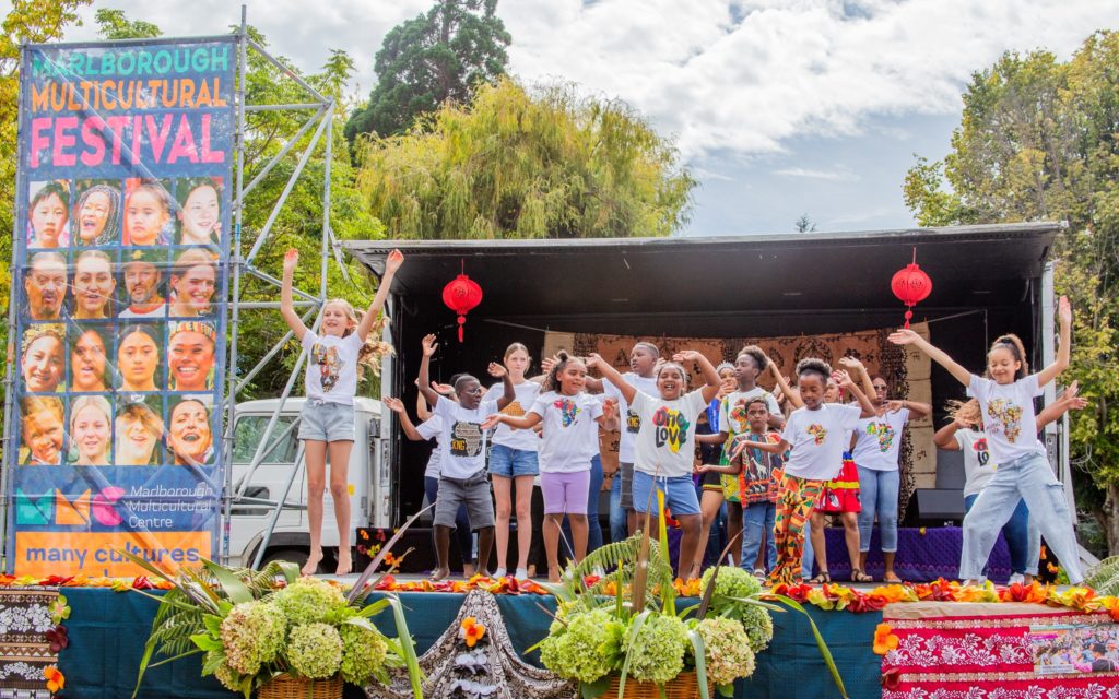 In this picture, taken at Marlborough Multicultural Festival 2025, a large group of pre-teen children perform on a crowded stage. The children are of mixed ethnicities and are wearing colourful t-shirts showing the African continent. The children appear happy, with many waving their hands in the air and smiling. To the left of the stage, a large banner displays smiling, happy faces of people of a variety of ethnicities beneath the title "Marlborough Multicultural Festival"