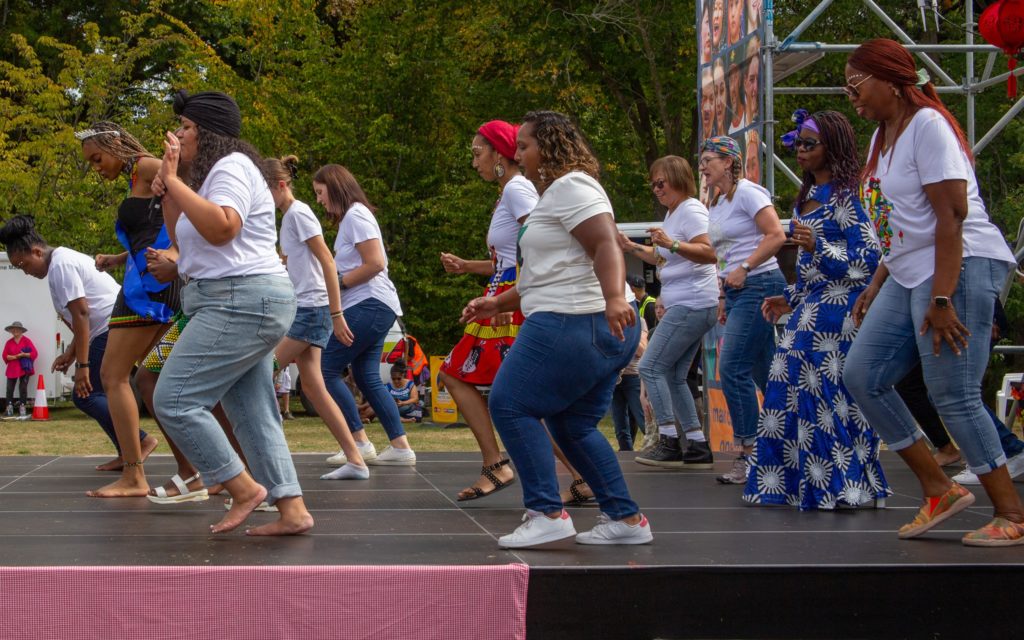 In this picture, taken at Marlborough Multicultural Festival 2025, a large group of women dance in lines on a stage. They are of various cultures and ethnicities and are dressed casually or in clothing of traditional African print. The women are pictured from the side, and all are stepping forward at the same time.
