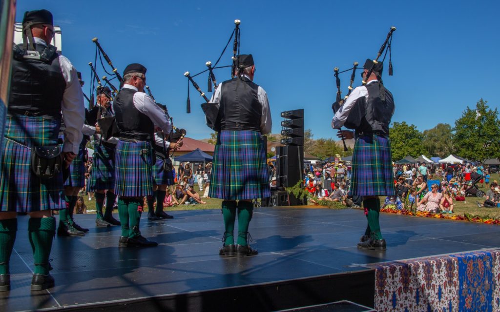 In this picture, taken at Marlborough Multicultural Festival 2025, a group of people are pictured from behind and playing bagpipes for a large audience who are seated on the grass in front of the stage. The people playing the bagpipes are wearing blue and green tartan kilts, long green socks and black waistcoats over long-sleeve white shirts. They are also wearing Scottish Balmoral hats.