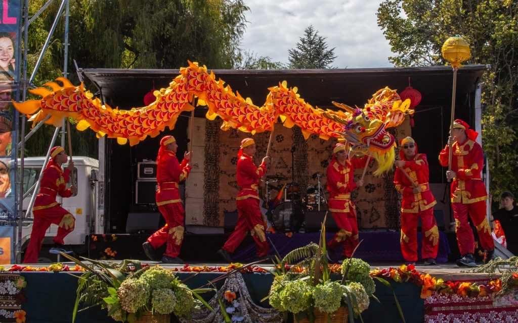 Taken at Marlborough Multicultural Festival 2025, a group of men dressed in red and gold traditional Chinese long-sleeved tunic suits are pictured performing on the stage. The men parade a red and gold Chinese dragon, which is held above their heads on long sticks. They men - and the dragon - are pictured in motion, parading on the stage.