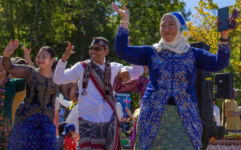 In this picture, taken at Marlborough Multicultural Festival 2025, two women and a man are seen in brightly coloured ethnic dress. They are facing the camera with both hands raised. The woman closest to the camera wears a heard scarf while all three are smiling.