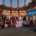Taken indoors, a large group of people stand in a semi-circle to be photographed. Those in front sit or kneel and behind them, others are standing. The people are of very diverse ethnicities, some in cultural dress, and Margaret Western is pictured standing in the centre of the group. Behind the group, a large digital screen says "Welcome to Marlborough District Council".