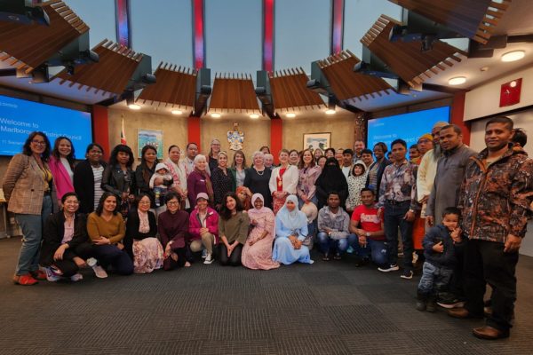 Taken indoors, a large group of people stand in a semi-circle to be photographed. Those in front sit or kneel and behind them, others are standing. The people are of very diverse ethnicities, some in cultural dress, and Margaret Western is pictured standing in the centre of the group. Behind the group, a large digital screen says "Welcome to Marlborough District Council".