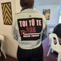 A woman, who is faced away, wears a denim jacket on the back of which is a black patch square. In it, red and white writing says, " Toi Tū Te Ora Rangatahi Symposium" where Te Whakaoreore was used.