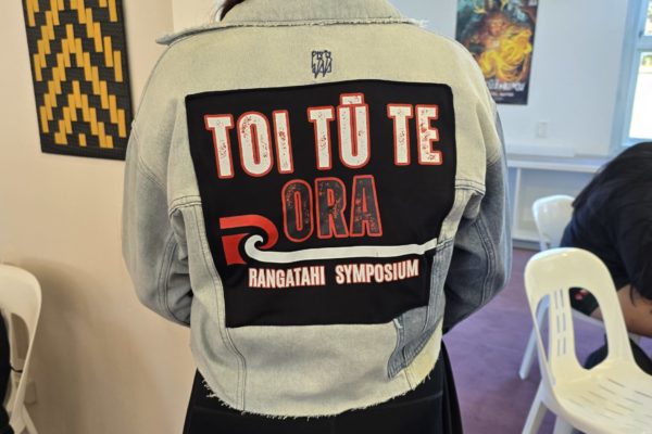 A woman, who is faced away, wears a denim jacket on the back of which is a black patch square. In it, red and white writing says, " Toi Tū Te Ora Rangatahi Symposium" where Te Whakaoreore was used.